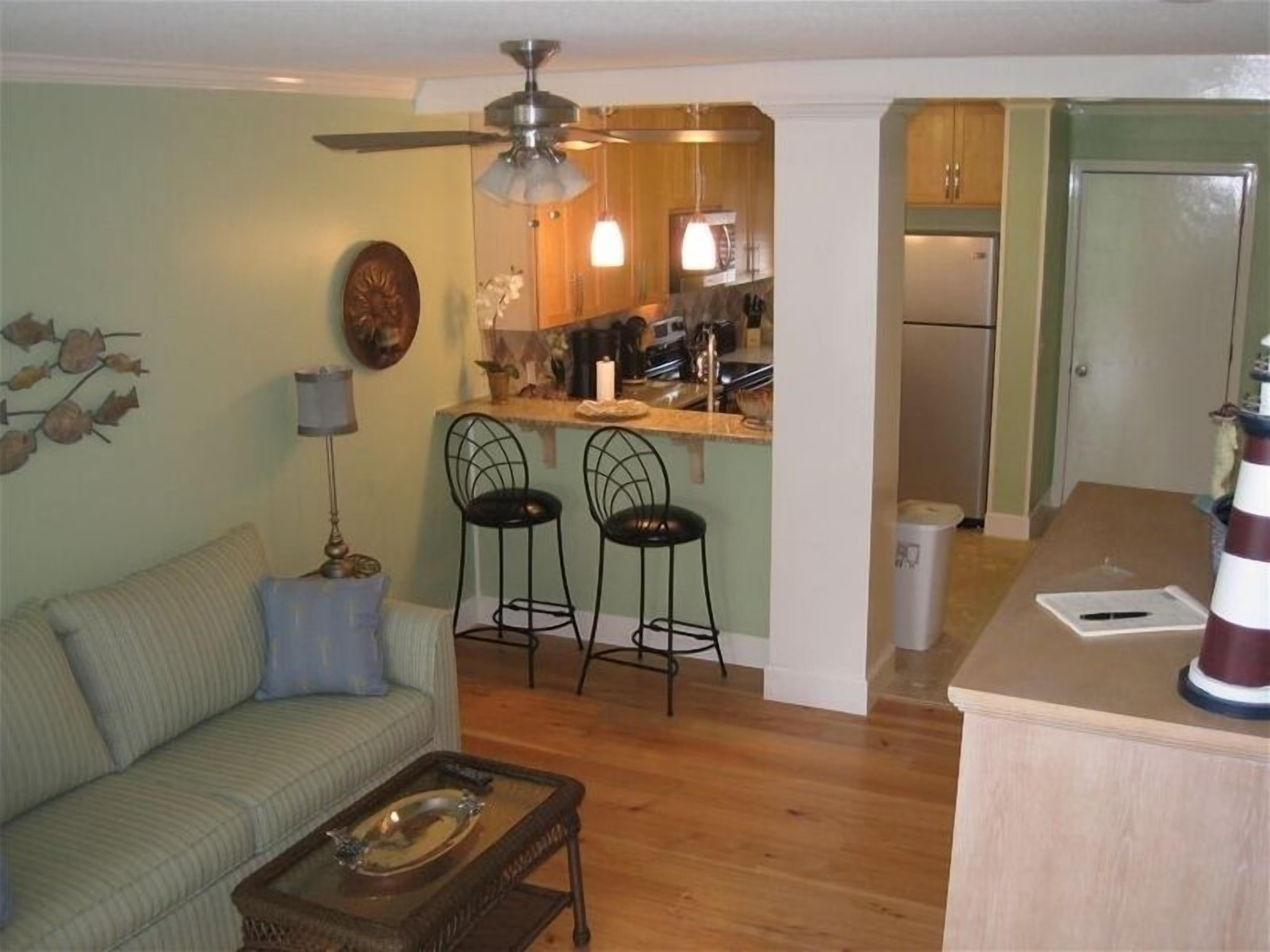 A living room with a sofa, two bar stools, a coffee table, and a kitchen area with a refrigerator and a sink.