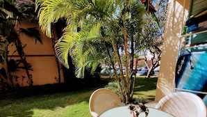 En udendørs terrasse med et rundt bord og hvide fletstole, omgivet af frodigt grønt og en klar blå himmel.