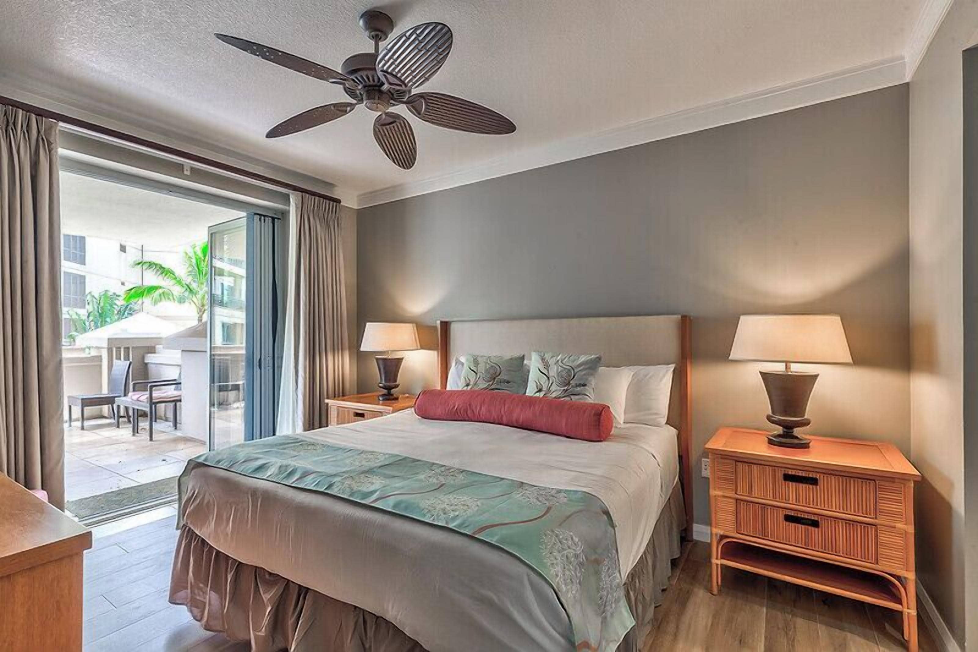 Une chambre d’hôtel comprenant un lit, deux tables de chevet avec des lampes, un ventilateur de plafond et une porte-fenêtre coulissante donnant sur un balcon.