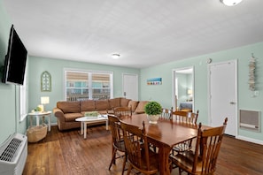 A living room with a brown couch, wooden dining table, and chairs, a television, and a window with blinds.