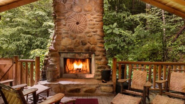 A stone fireplace with a fire, surrounded by wooden furniture and a wooden deck. - RIVERFRONT LUXURY LODGE/4 KING SUITES/Swim Spa/Game Room/Fire Pit/4,200 Sq. Ft. (Sevierville)