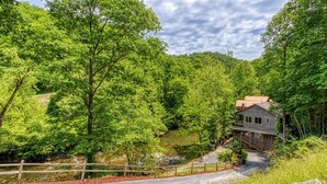 A rustic cabin with a stone exterior, surrounded by lush greenery and a small stream. - RIVERFRONT LUXURY LODGE/4 KING SUITES/Swim Spa/Game Room/Fire Pit/4,200 Sq. Ft. (Sevierville)