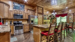 A wooden cabin-style kitchen with a central island, wooden cabinets, and a dining area with rustic chairs. - RIVERFRONT LUXURY LODGE/4 KING SUITES/Swim Spa/Game Room/Fire Pit/4,200 Sq. Ft. (Sevierville)