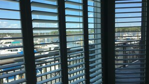 Une vue à travers les stores d'une fenêtre, donnant sur une marina où des bateaux sont amarrés.