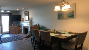 Un coin repas avec une table dressée pour quatre personnes, des chaises, un lustre, une cheminée et un tableau accroché au mur.