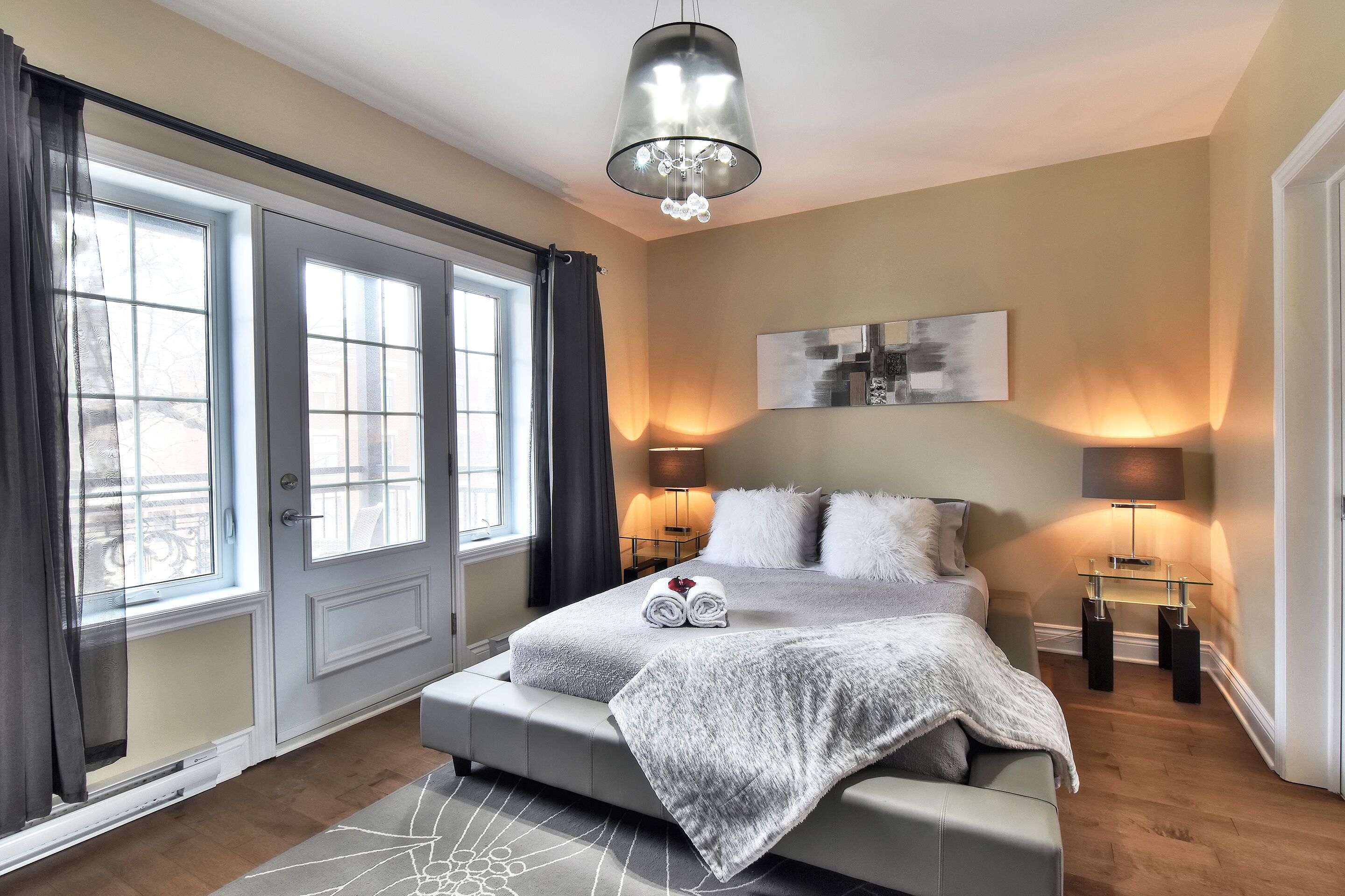 A modern bedroom with a large bed, two bedside lamps, a chandelier, and a view of the city through large windows.