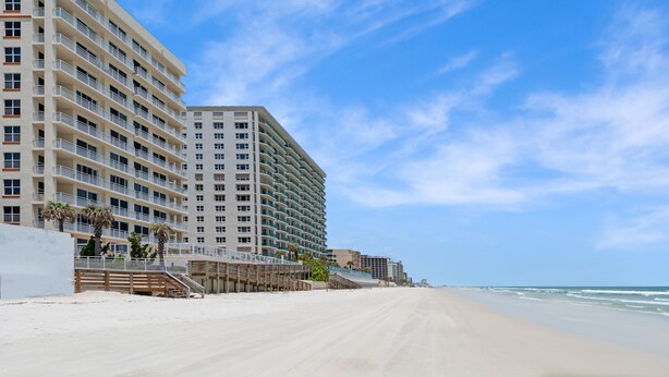Un hôtel situé en bord de mer, doté de nombreux balcons, d’une plage de sable et offrant une vue sur un ciel bleu azur.