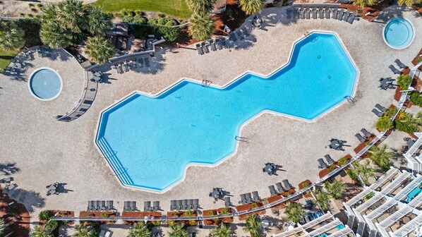 An aerial view of a resort with a large central swimming pool, multiple lounge areas, and surrounding greenery.