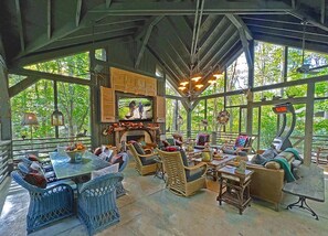 Un vaste salon extérieur aménagé avec des meubles en osier, une cheminée et de grandes fenêtres offrant une vue sur la verdure environnante.
