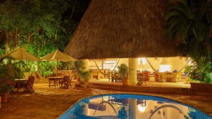 An indoor pool area with thatched umbrellas, wooden furniture, and lush greenery.