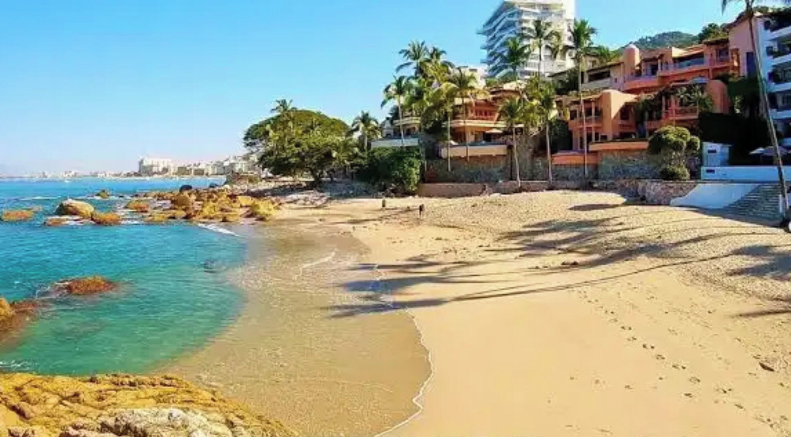 Une plage à l’eau turquoise et limpide, avec un rivage sablonneux et des bâtiments dotés de balcons donnant sur la mer.
