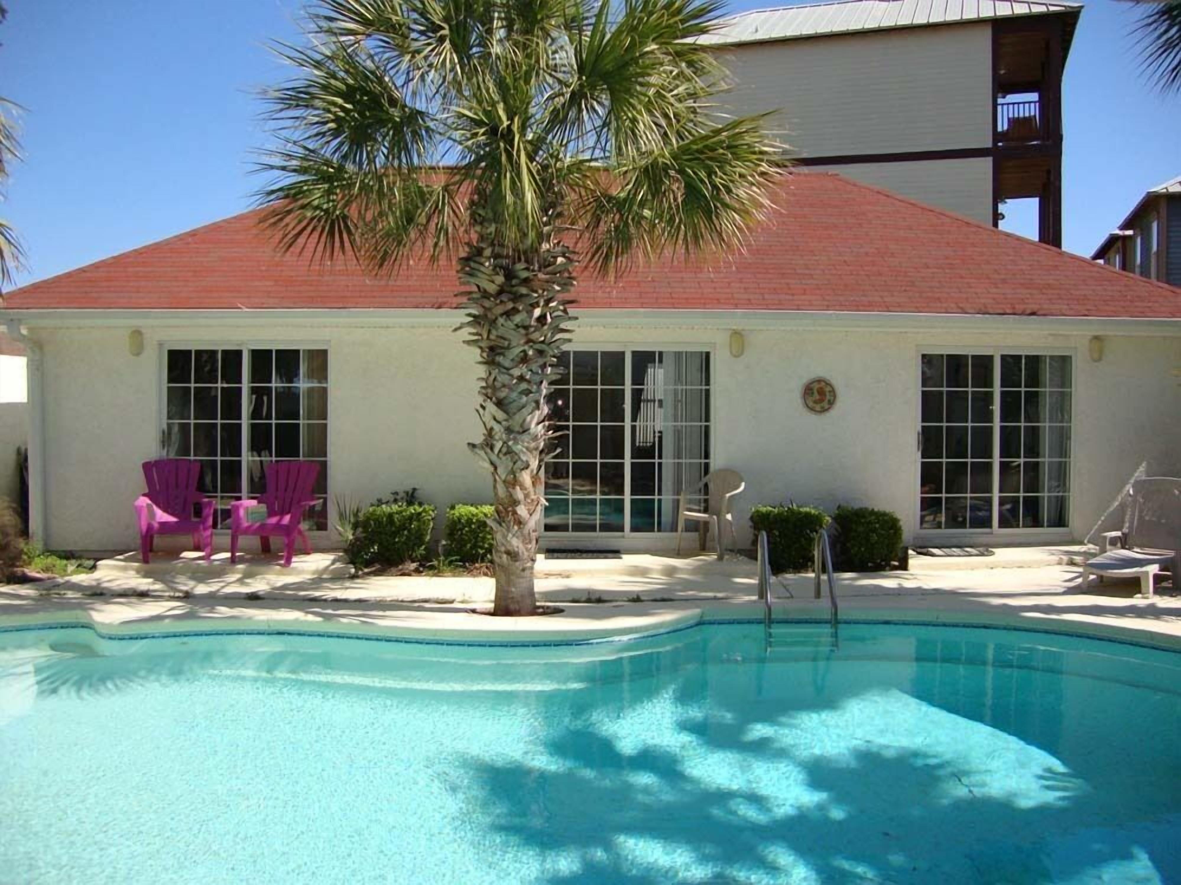 A pool area with a palm tree, two purple chairs, and a building with a red roof and large windows.