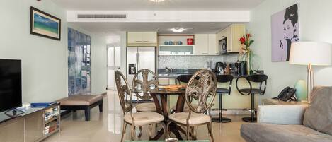 A modern living room with a flat-screen TV, a sofa, a dining table with chairs, and a kitchen area in the background.