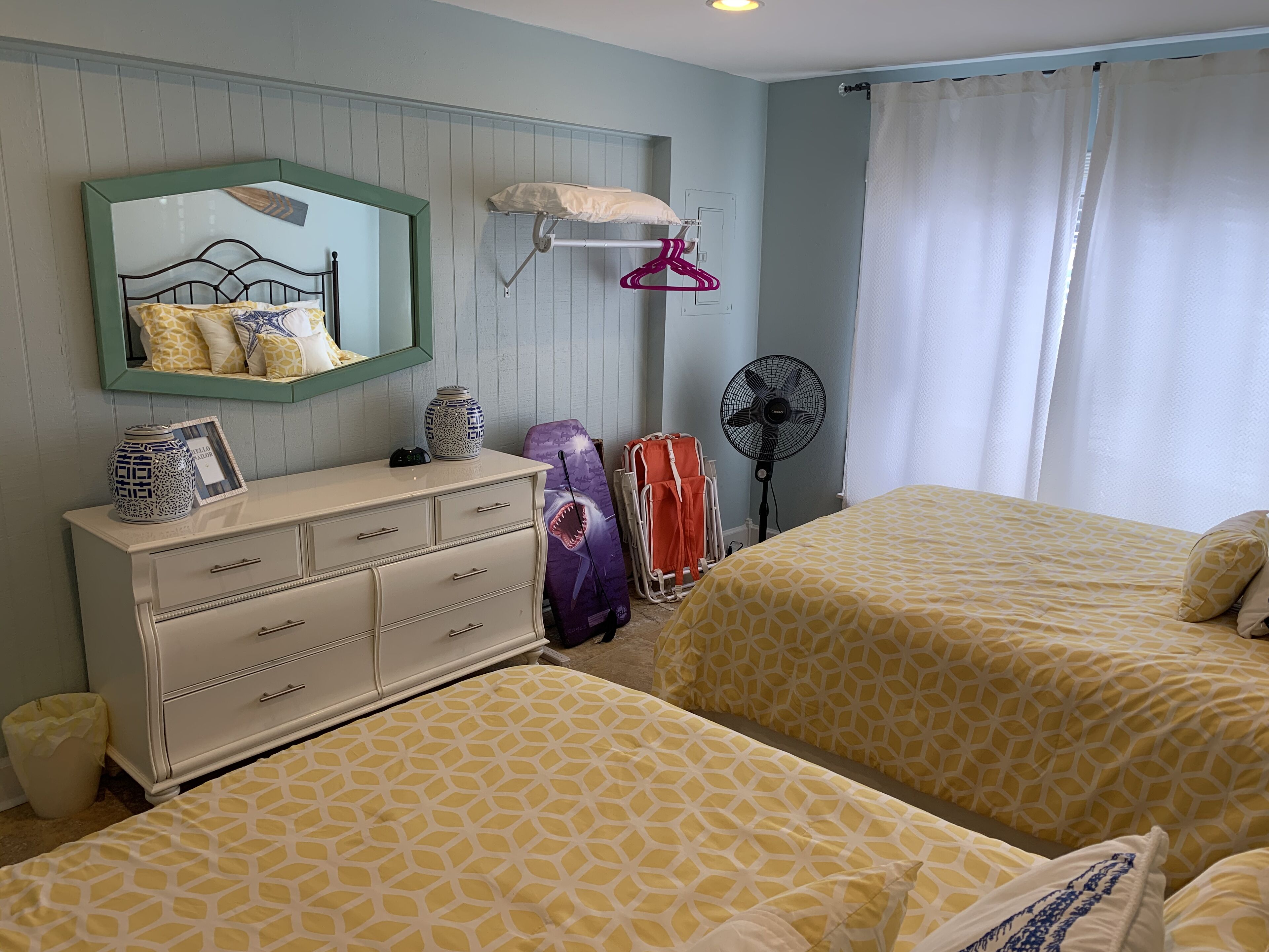 A bedroom with two beds, a dresser, a mirror, a fan, and a window with curtains.