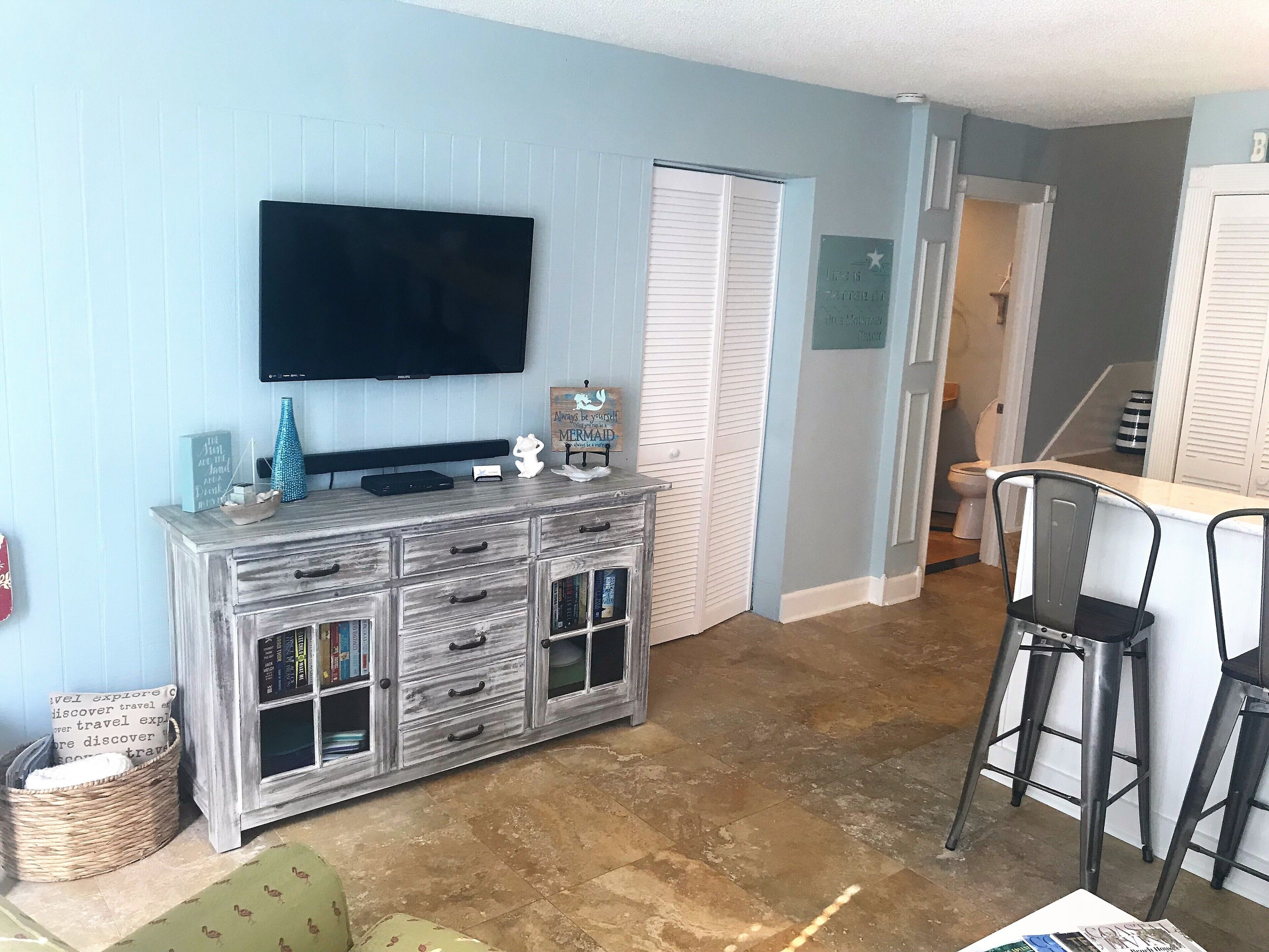 Una sala de estar con una televisión, un mueble de madera con cajones, una cesta de mimbre y una silla verde.