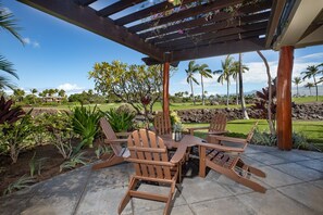 An outdoor patio with wooden Adirondack chairs and a table, surrounded by lush landscaping and palm trees.