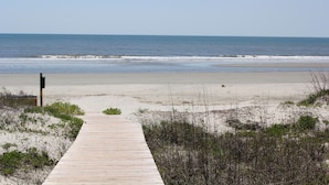 Ein Holzsteg führt zu einem Strand mit Dünen und spärlicher Vegetation.