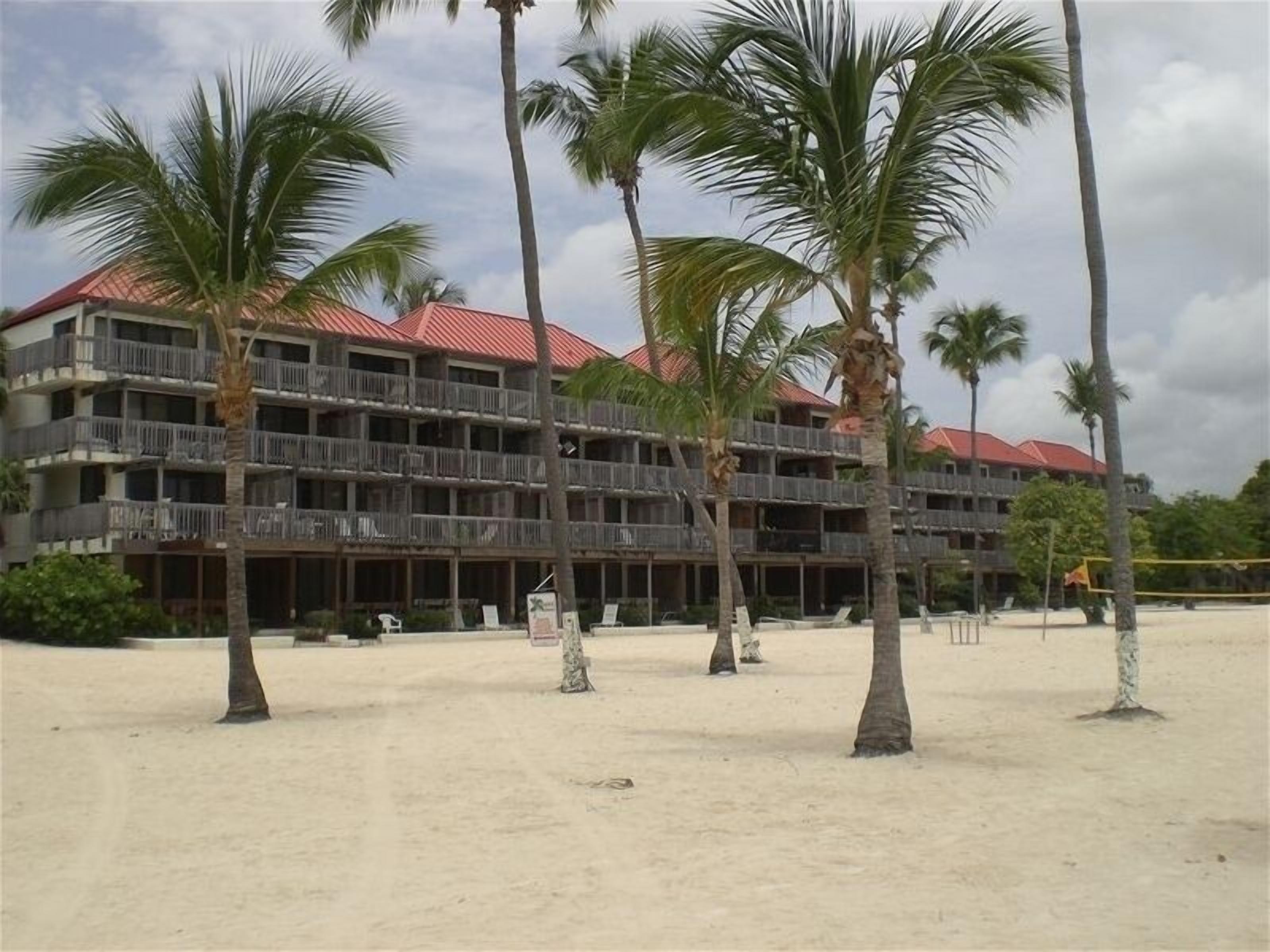 A multi-story hotel with balconies, located on a sandy beach with palm trees.