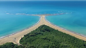 Vue aérienne d’une plage dont le rivage est incurvé, avec une forêt dense au premier plan et des eaux bleues et calmes.