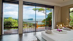 Quarto com uma grande porta de vidro deslizante que dá para uma varanda com vista para a água e vegetação.