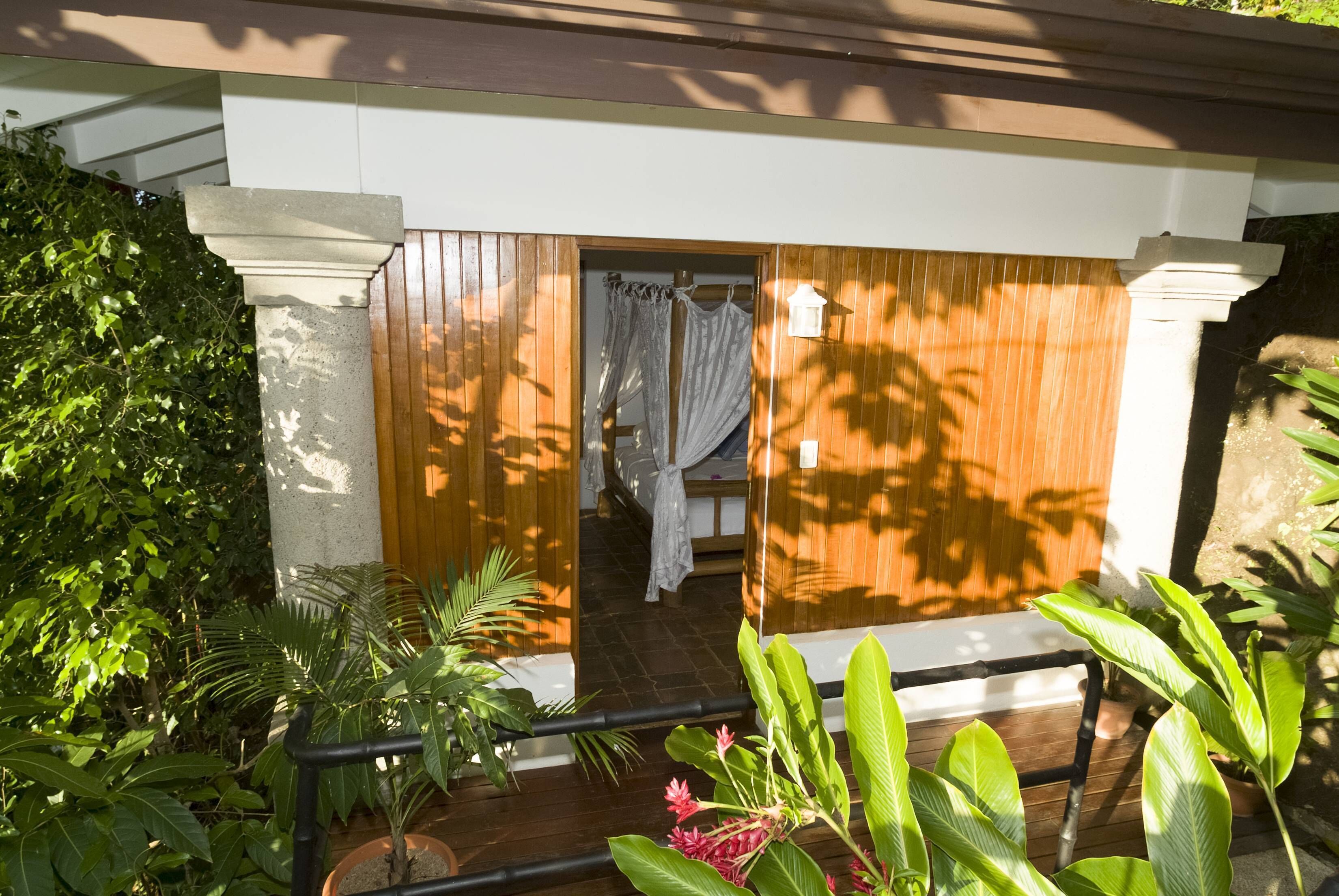Een houten terras met plantenpotten, een witte deur en een kamer met een bed en gordijnen.