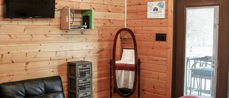 A wooden cabin room with a bed, a black leather chair, a television mounted on the wall, and a small cabinet with electronic devices.