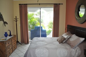Une chambre à coucher avec un lit, une commode en bois, une chaise bleue et une vue sur un balcon agrémenté de plantes.