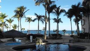 Área de piscina em resort, com palmeiras, estrutura de telhado de palha e vista para o oceano.