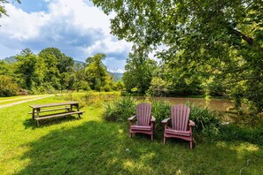 A picnic area with a wooden bench and two Adirondack chairs by a pond surrounded by lush greenery.