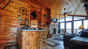A wooden cabin interior with a kitchen area, a dining table with chairs, a sofa, and a TV mounted on the wall.