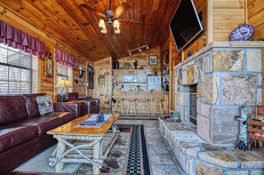 A cozy cabin interior with a wooden ceiling, stone fireplace, leather sofa, and a dining area.