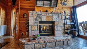 A cozy cabin interior with a stone fireplace, wooden walls, a mounted TV, and a ladder to the right.