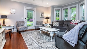 A living room with a dark grey sofa, two armchairs, a white coffee table, and a patterned rug. There are two lamps on side tables, a ceiling fan, and a clock on the wall.