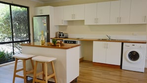 A kitchen with white cabinets, a wooden countertop, a built-in dishwasher, and a washing machine. There are two stools and a bottle of alcohol on the counter. - Farm Escape - Spacious Cottage in Kangaroo Valley (Kangaroo Valley)