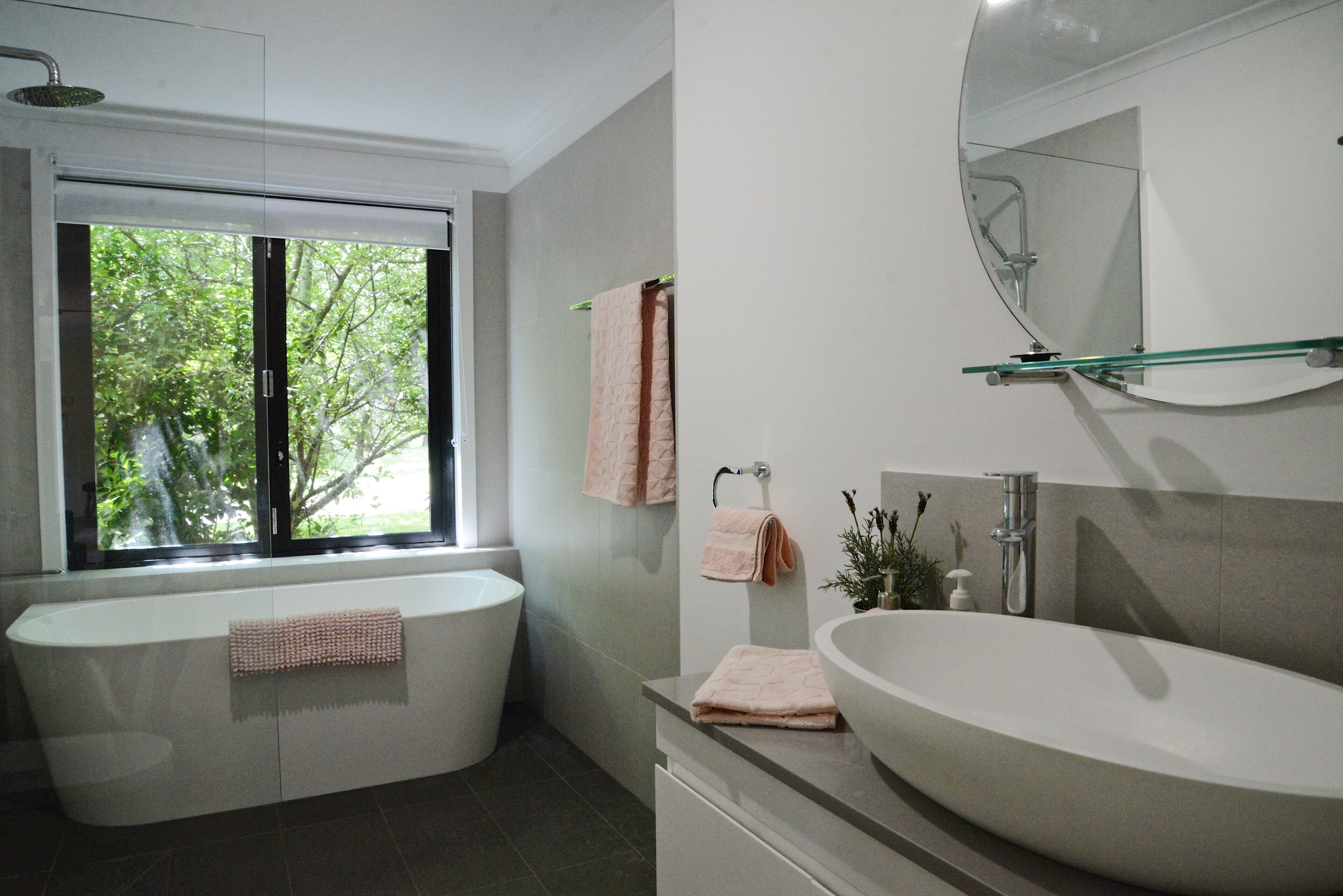 Ein modernes Badezimmer mit einer freistehenden Badewanne, einem großen Waschtisch mit rundem Spiegel und einem Fenster mit Blick auf Grünflächen.