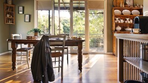 A dining area with a wooden table and chairs, a coat hanging on one chair, and a view through a glass door to the outdoors. - Kookaburra Ridge - beautiful home set on a 1.6 acre garden with fireplace & spa (Hepburn)