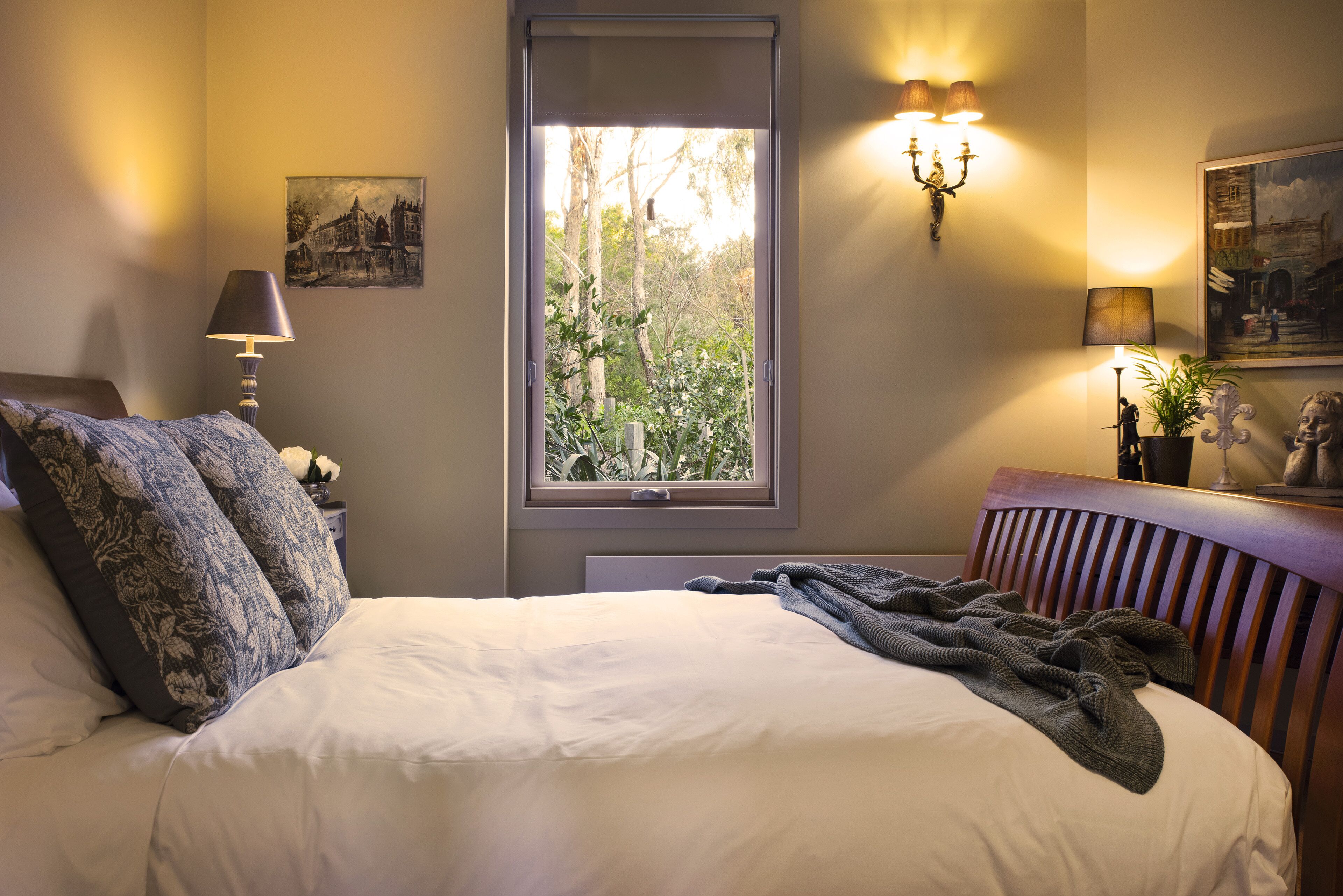 A neatly made bed with a white duvet and a grey blanket, a bedside lamp, a framed picture on the wall, and a window with a view of trees.