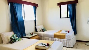 Quarto de hotel com duas camas, cada uma com colcha branca e manta dourada. Cortinas azuis nas janelas, e o chão do quarto é coberto por azulejos.