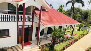 Um edifício de dois andares com telhado vermelho, varanda coberta e um pequeno jardim com vasos de plantas e uma palmeira.
