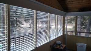 A room with large windows covered by blinds, a wooden ceiling, and a small table with a trash bin.