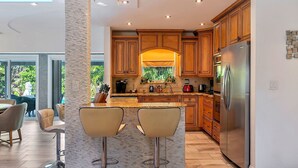 A modern kitchen with wooden cabinets, a stainless steel refrigerator, and a bar area with two stools. - Renovated 3BR/2BA with large heated saltwater pool & tiki bar (Oakland Park)