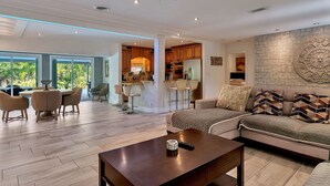 A spacious living area with a sofa, coffee table, and dining set. A kitchen with modern appliances and a stone accent wall is visible in the background. - Renovated 3BR/2BA with large heated saltwater pool & tiki bar (Oakland Park)