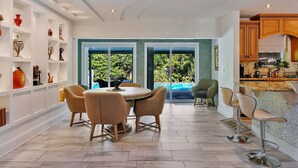A spacious dining area with a round table and chairs, a kitchen with wooden cabinets, and large windows offering a view of greenery. - Renovated 3BR/2BA with large heated saltwater pool & tiki bar (Oakland Park)