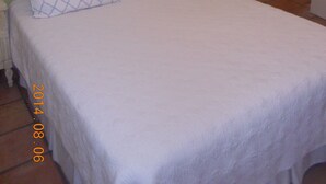 Valkoinen pääty ja kuviollinen tyyny koristavat sänkyä, sen vieressä on pieni yöpöytä, jossa on lamppu, ja seinällä on taulu.
