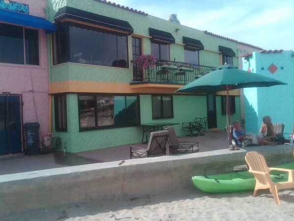 A two-story building with a balcony, outdoor seating, and a green kayak on the sand.