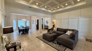 A spacious living room with a large sectional sofa, a dining area with a chandelier, and a kitchen in the background.