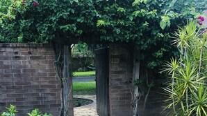 A brick pathway leading to a wooden gate, surrounded by lush greenery and flowering plants.