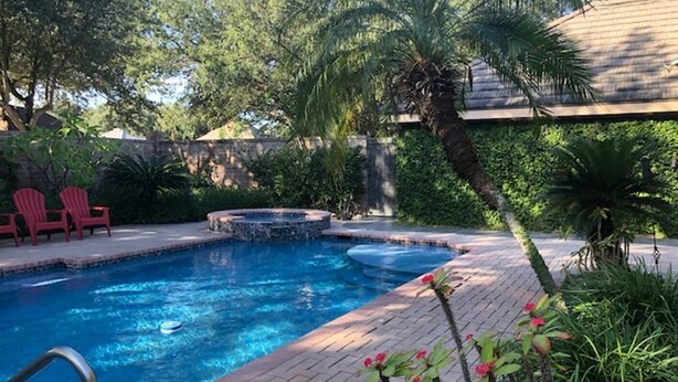 A swimming pool with a hot tub, surrounded by a brick patio, palm trees, and red chairs.