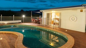 Una piscina con muelle de madera, un porche cubierto con mobiliario exterior y un edificio blanco con una puerta.