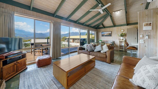 Amplia sala de estar con televisor grande, muebles de madera, ventilador de techo y vistas a montañas a través de amplios ventanales.